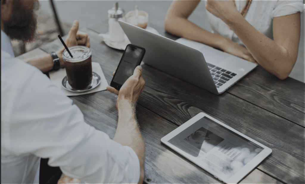 Imagem mostra de perto uma mesa marrom, do lado esquerdo um homem com um celular na mão e um copo de bebida com canudinho em sua frente, e do lado direito da imagem aparece os braços de uma mulher com um notebook em sua frente.