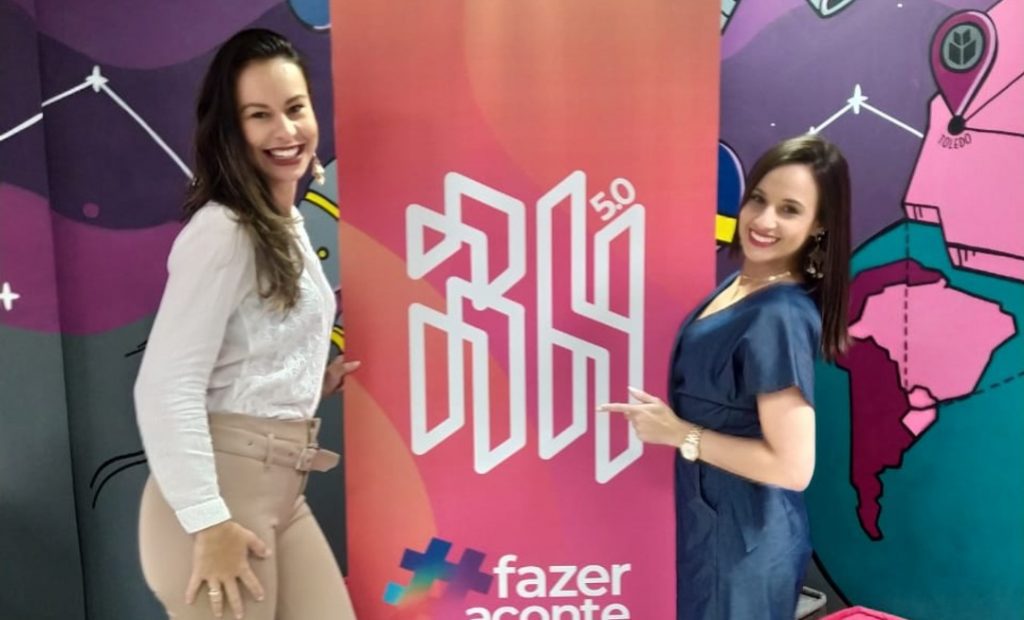 Imagem mostra duas mulheres de pé sorrindo, e entre as mulheres contém um banner rosa com laranja e no centro uma Logo do Workshop Tech Recruiter.