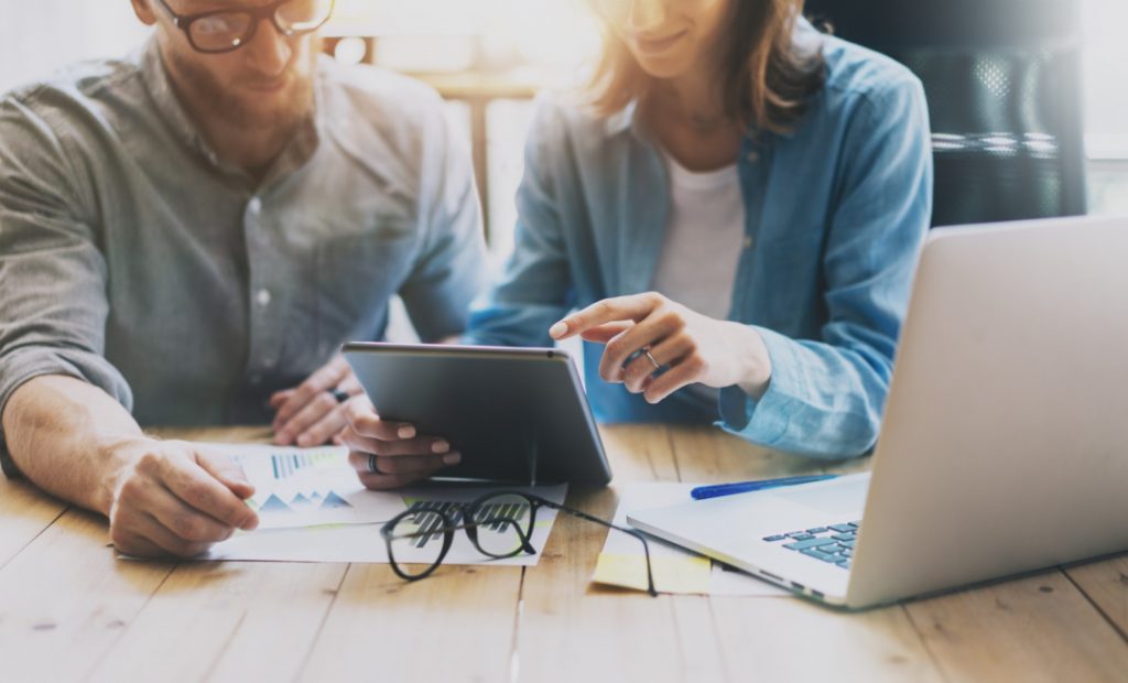 Imagem mostra um homem e uma mulher sentados de frente para uma mesa de escritório, com um notebook sobre a mesa. Do lado esquerdo esta o homem e do do lado direito esta a mulher segurando um tablet.