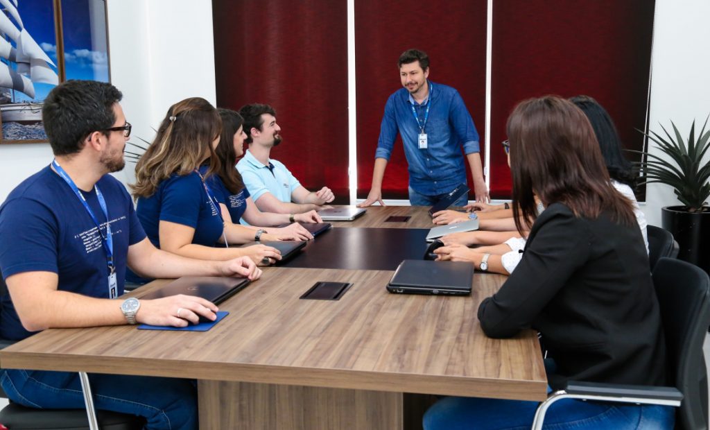 Imagem mostra uma sala de reunião, com um homem em pé em frente a mesa, quatro pessoas sentadas do lado esquerdo da mesa e outras três pessoas sentadas do lado direito.