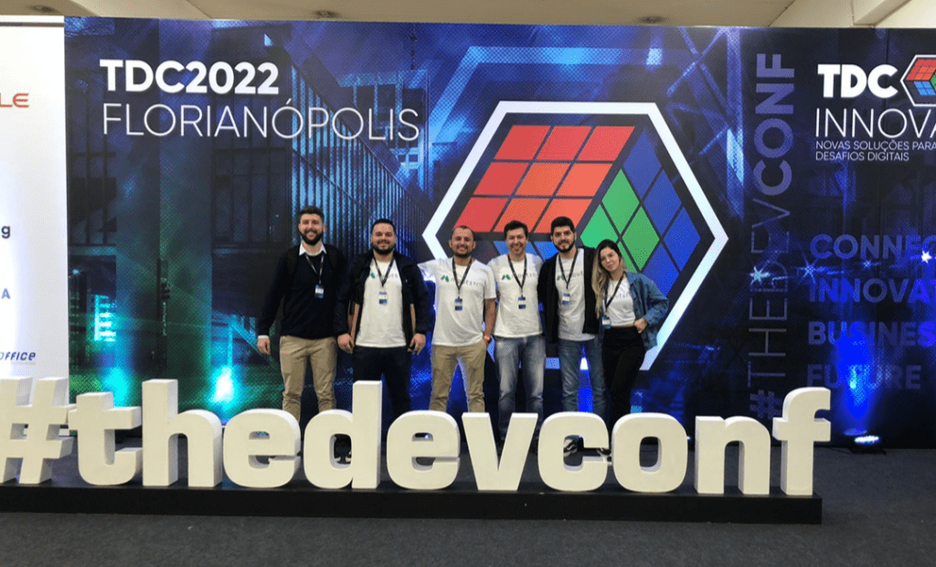 Imagem mostra cinco homens e uma mulher juntos, com uma frase na frente dizendo #thedevconf. No fundo contém um banner com o desenho de um cubo mágico, com uma escrita no canto superior esquerdo dizendo TDC2022 Florianópolis.