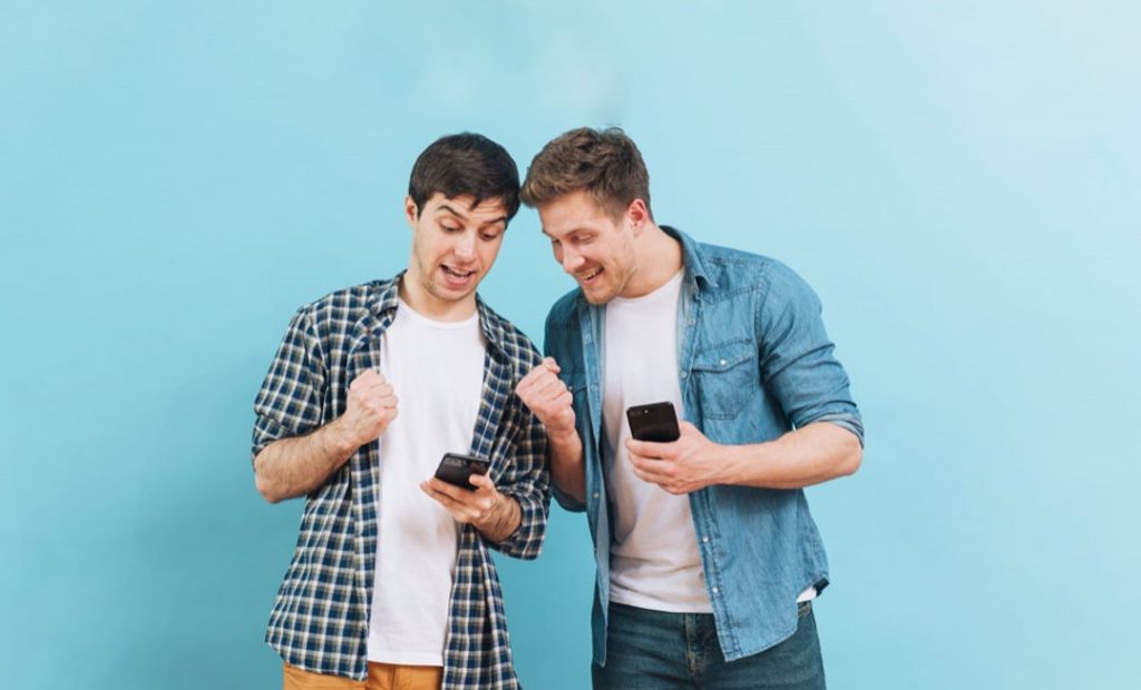 Foto de dois homens em pé segurando seus celulares e comemorando enquanto olham para um dos celulares. Atras deles em segundo plano tem uma parece de cor azul claro