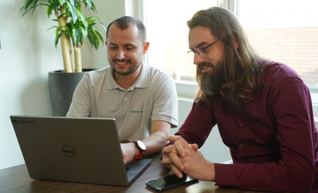 Foto de escritório com dois homens sentados olhando para um notebook apoiado sobre uma mesa.