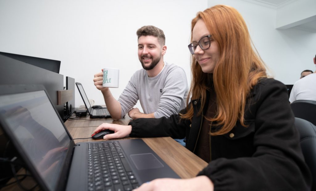 Foto de uma homem e uma mulher sentados em uma mesa de escritório e um notebook sobre a mesa. O Homem toma café olhando para a tela do notebook enquanto a mulher movimenta o mouse