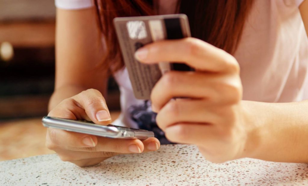 Foto de uma mulher segurando um celular na mão direita e um cartão de crédito na mão esquerda. Parece estar realizando uma compra online.