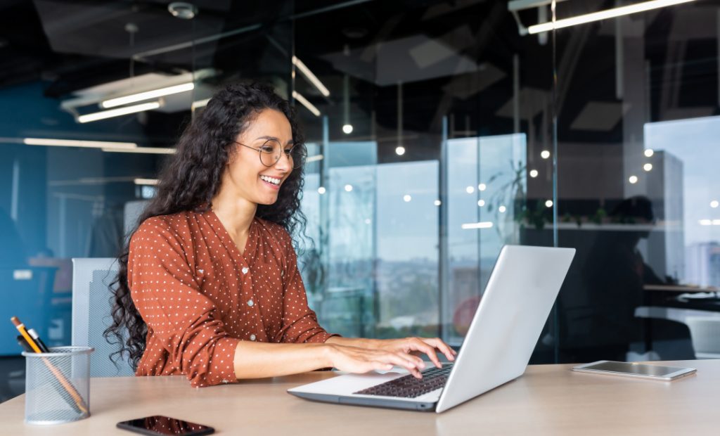 Foto de escritório, mulher sentada utilizando um notebook. A mulher esta sorrindo enquanto digita algo