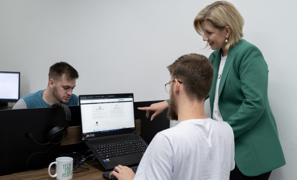 Foto de ambiente de escritório com dois homens sentados usando notebook, um dos homens esta sendo orientado por uma mulher que aponta para a tela do notebook enquanto conversa com ele