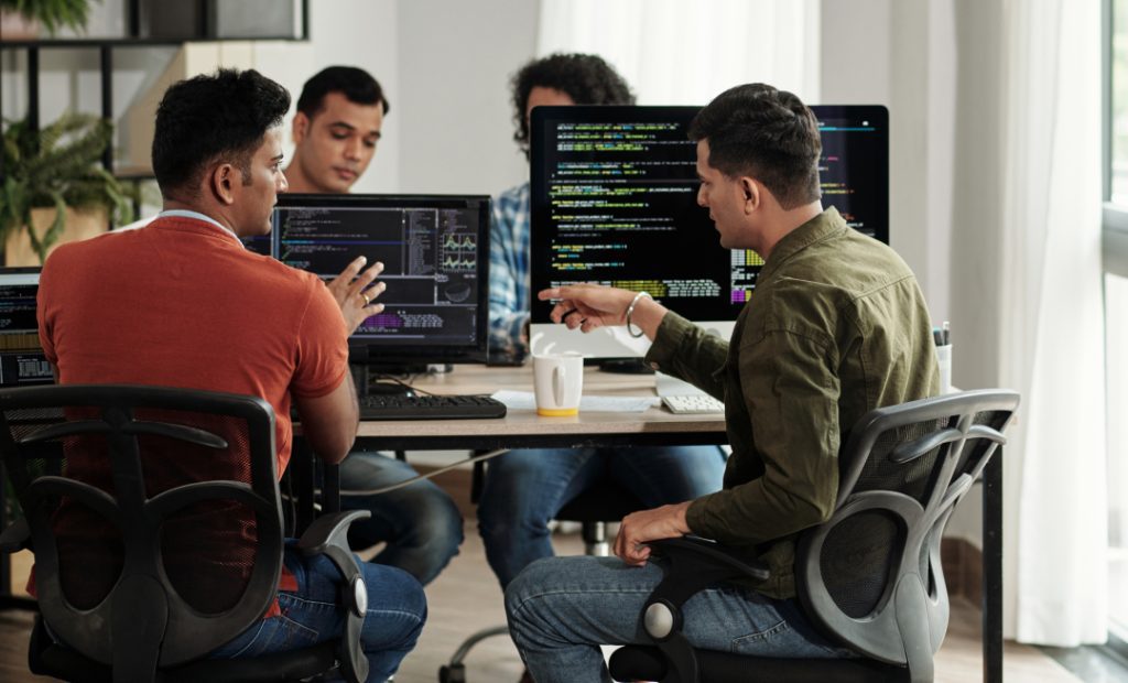 Imagem de escritório com uma mesa central onde tem quatro desenvolvedores sentados trabalhando e conversando. Sob a mesa tem computadores e monitores mostrando dados aleatórios. Do lado direito da foto é possível ver a luz do dia entrando pela janela