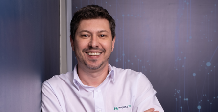 Foto de um homem sorrindo para a câmera. O homem esta com uma camisa branca encostado na parece posando para a foto onde parece ser um escritório.