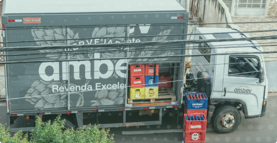 Imagem mostra caminhão de bebidas estacionado, com uma de suas portas laterais abertas, ao lado do caminhão contém duas pilhas de caixas de cerveja.