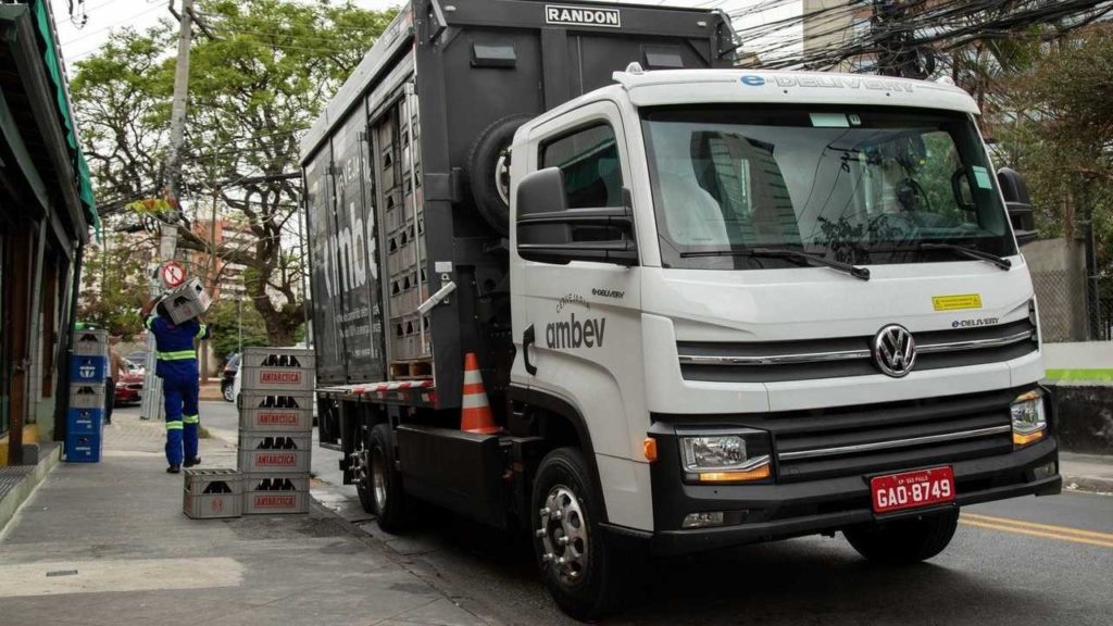 Imagem mostra um caminhão de carga da estacionado em frente a um comércio, ao laldo do caminhão esta um homem de pé e uma pilha de caixas de cerveja.