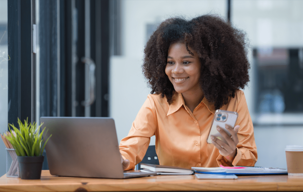 Imagem mostra uma mulher em uma sala de escritória, ela esta sorrindo, sentada diante uma mesa, com um notebook em sua frente.